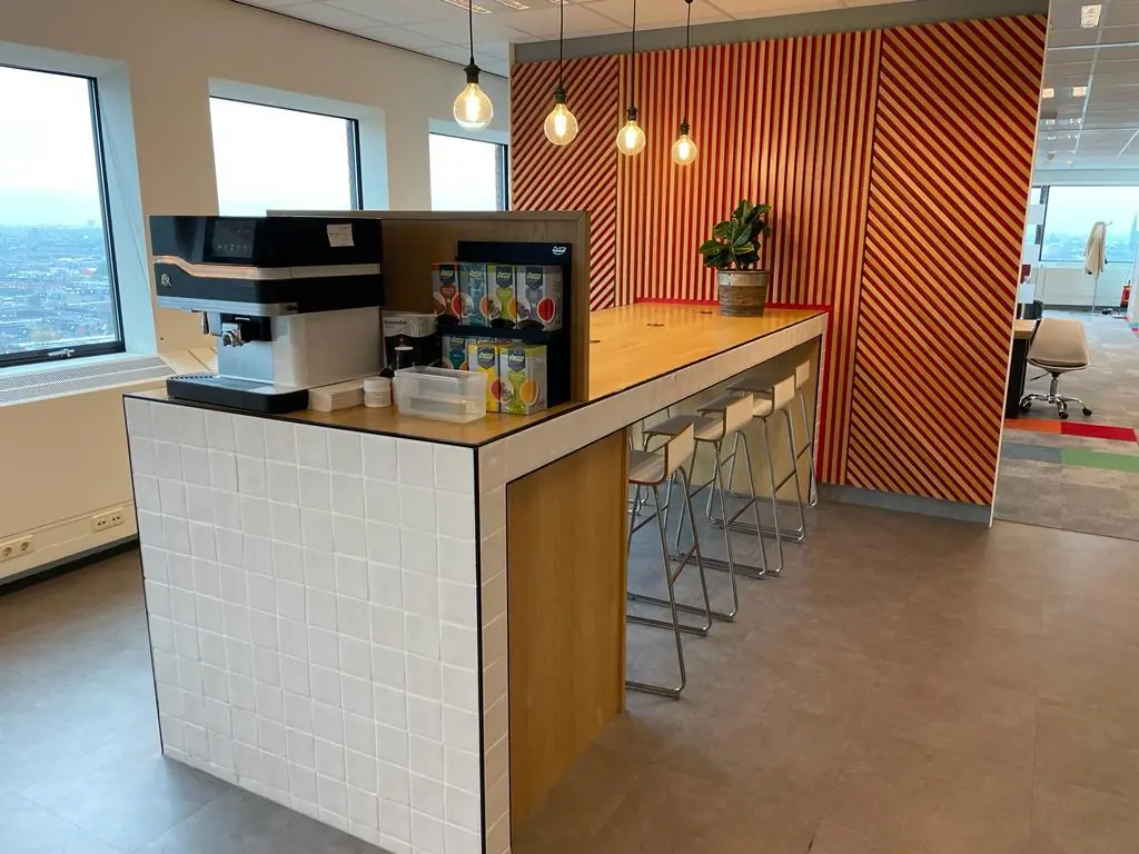 Moderne koffiehoek met een lange houten tafel, barkrukken en een koffieautomaat op het Johanna Westerdijkplein.